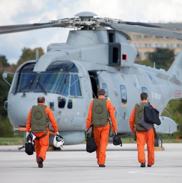 3 male pilots, dressed in orange flight suits and holding white helmets, walk towards an AW101 Merlin helicopter, with their backs turned to the camera