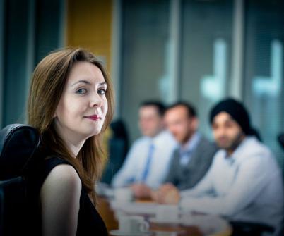 Woman looks over her shoulder while sitting at boardroom table. Three men sit at opposite side of table, out of focus.