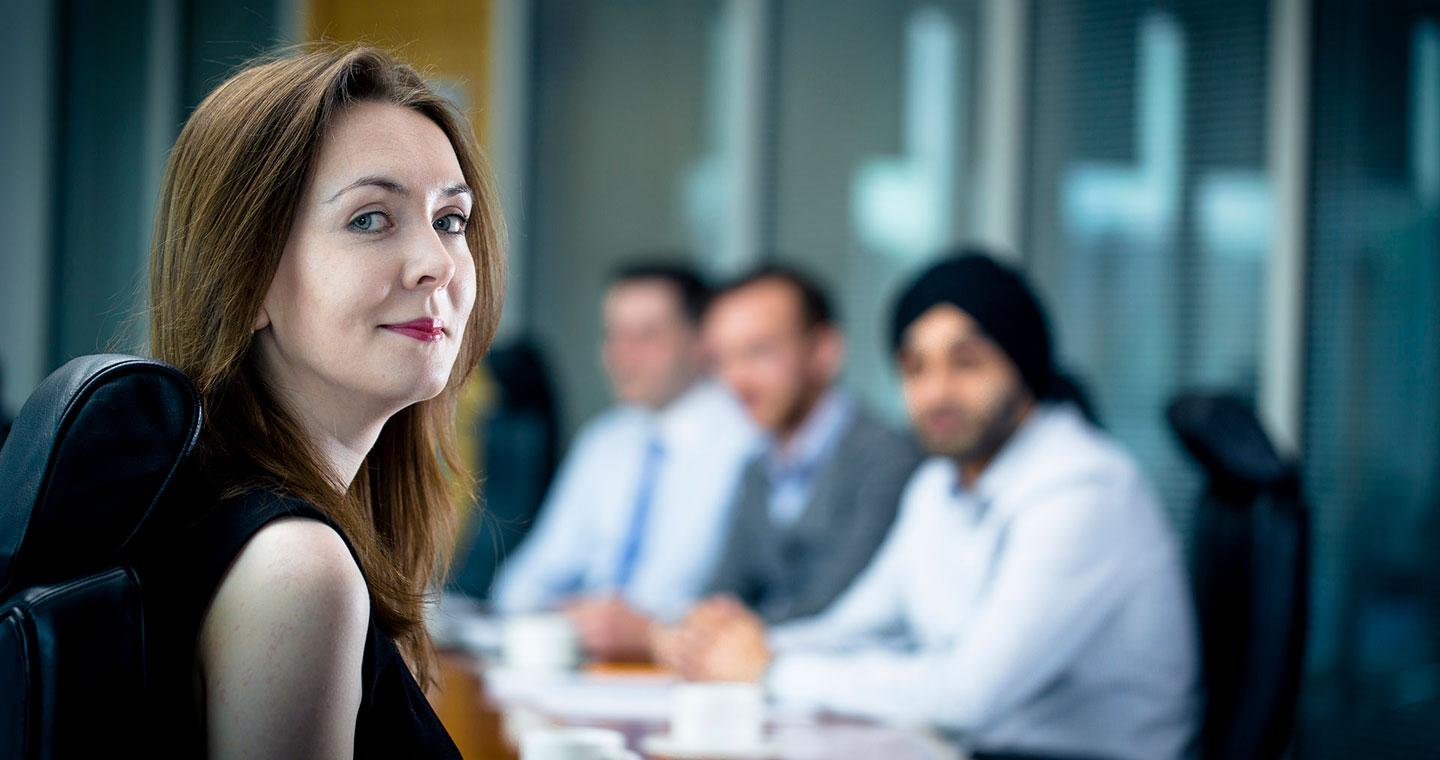 Woman looking over shoulder and smiling at boardroom table, with three men at other side of table, out of focus