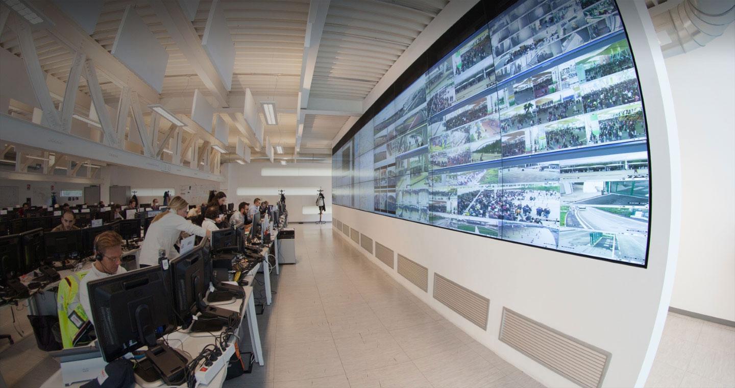 Operations room with desks of people working facing a giant wall of screens that are showing flows of people around a major event