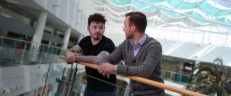 Two young men, one in a t shirt with tattoos, one in a shirt and cardigan, lean on balcony overlooking atrium at Leonardo site