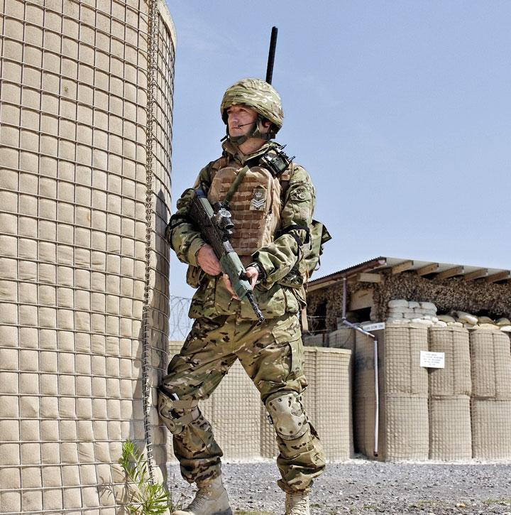 Soldier in full camouflage, holding rifle, leans on a wall and looks into the distance