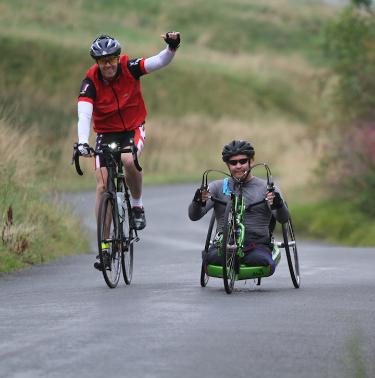 Cyclist with arm raised riding alongside a disabled rider on a customised 3 wheel bike