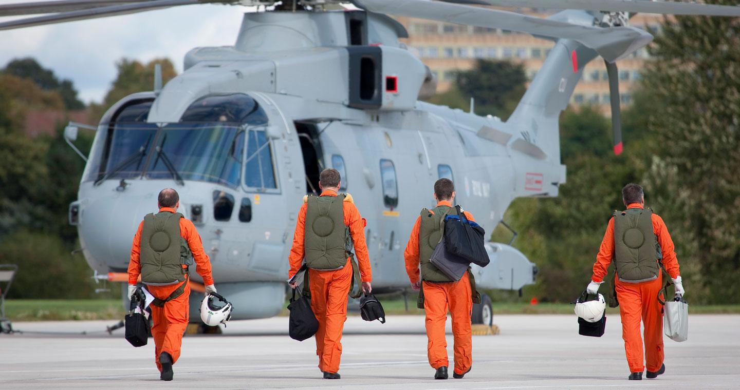 4 male pilots, dressed in orange flight suits and holding white helmets, walk towards an AW101 Merlin helicopter, with their backs turned to the camera